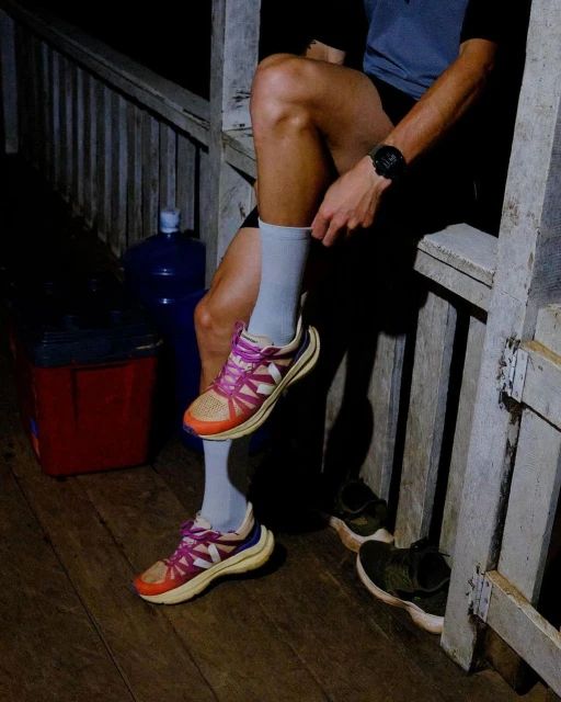 An adult wearing grey socks and colorful sneakers sits on a porch with wooden railings next to water bottles and other athletic shoes.