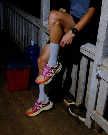 An adult wearing grey socks and colorful sneakers sits on a porch with wooden railings next to water bottles and other athletic shoes.