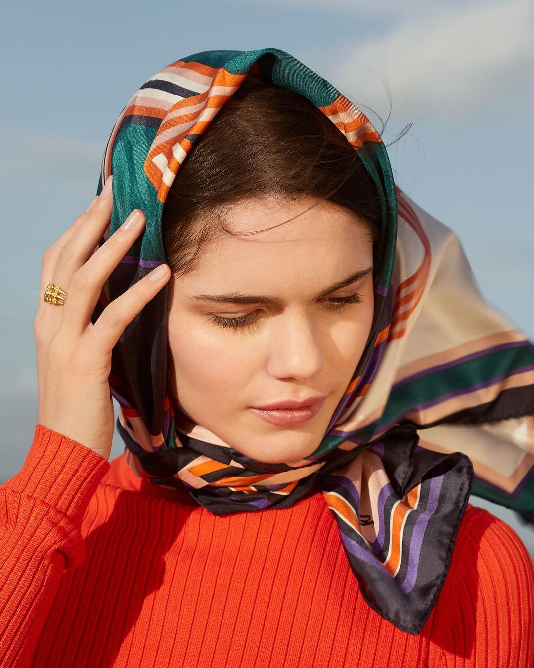 Woman in orange top and patterned headscarf poses with hand near face in an eye-level shot against a blurred, bright-blue background.