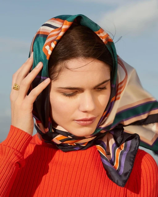 Woman in orange top and patterned headscarf poses with hand near face in an eye-level shot against a blurred, bright-blue background.