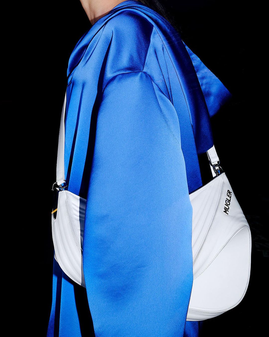 A model wears a vivid blue satin hooded top and a white Mugler shoulder bag against a dark backdrop in a fashion-forward studio shot.