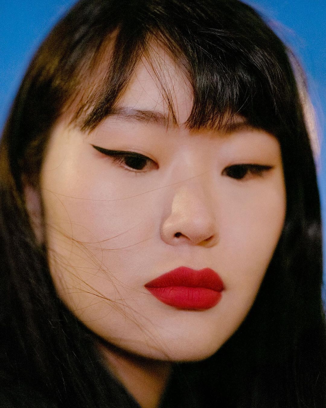 Close-up portrait of an Asian woman with striking makeup, including red lipstick and winged eyeliner, against a soft blue background.