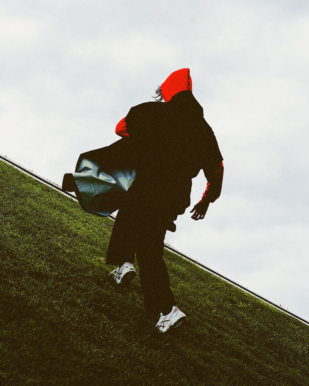 Person in a red hooded sweatshirt and black pants running uphill on a grassy hill against a cloudy sky, carrying a bag.
