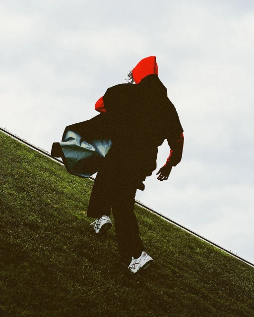 Person in a red hooded sweatshirt and black pants running uphill on a grassy hill against a cloudy sky, carrying a bag.