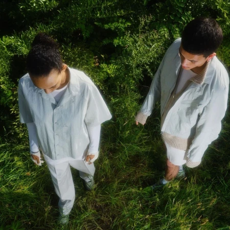 High-angle shot of two models walking through lush green grass, wearing light grey jackets and white trousers.