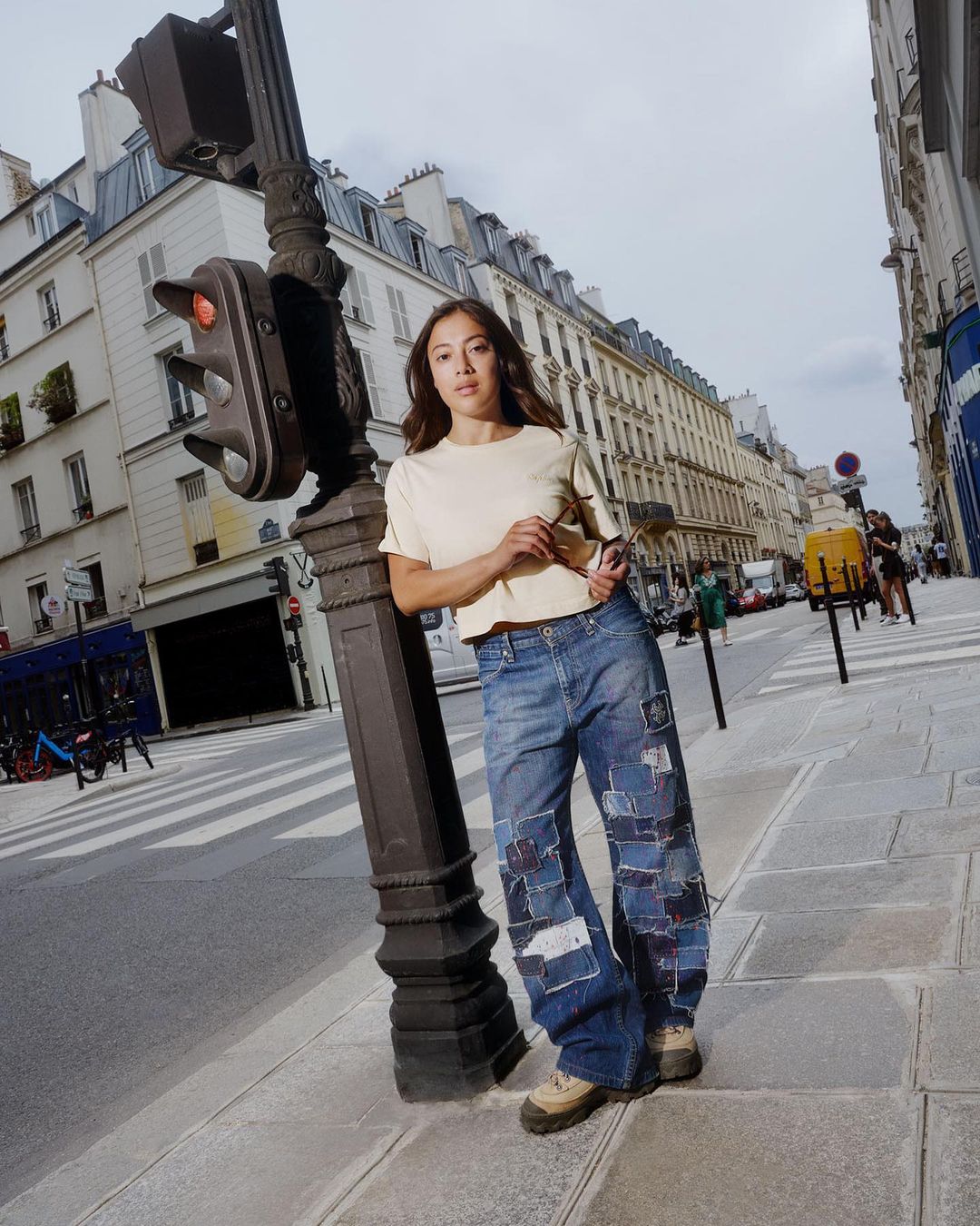 Woman in jeans and a t-shirt posing by a traffic light on an urban street in Paris, France, with buildings and crosswalk in the background.