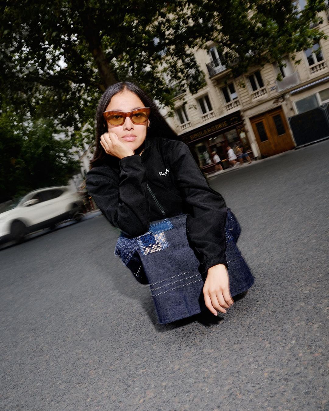 Woman in sunglasses and jacket poses on asphalt in an urban setting with a dark denim bag, with a blurred car and buildings visible behind.