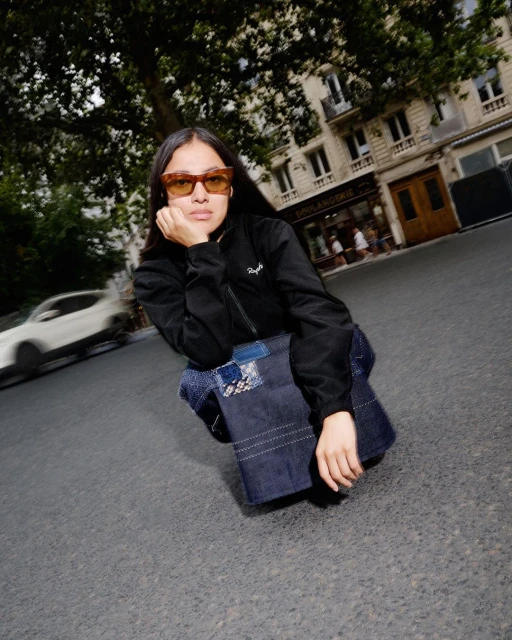 Woman in sunglasses and jacket poses on asphalt in an urban setting with a dark denim bag, with a blurred car and buildings visible behind.