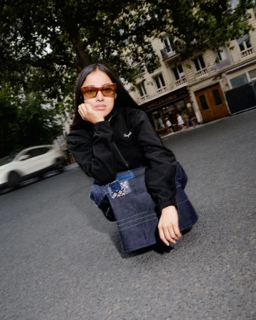 Woman in sunglasses and jacket poses on asphalt in an urban setting with a dark denim bag, with a blurred car and buildings visible behind.