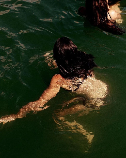 Two women are swimming in dark green water, with ripples and sunlight creating a natural and serene scene.
