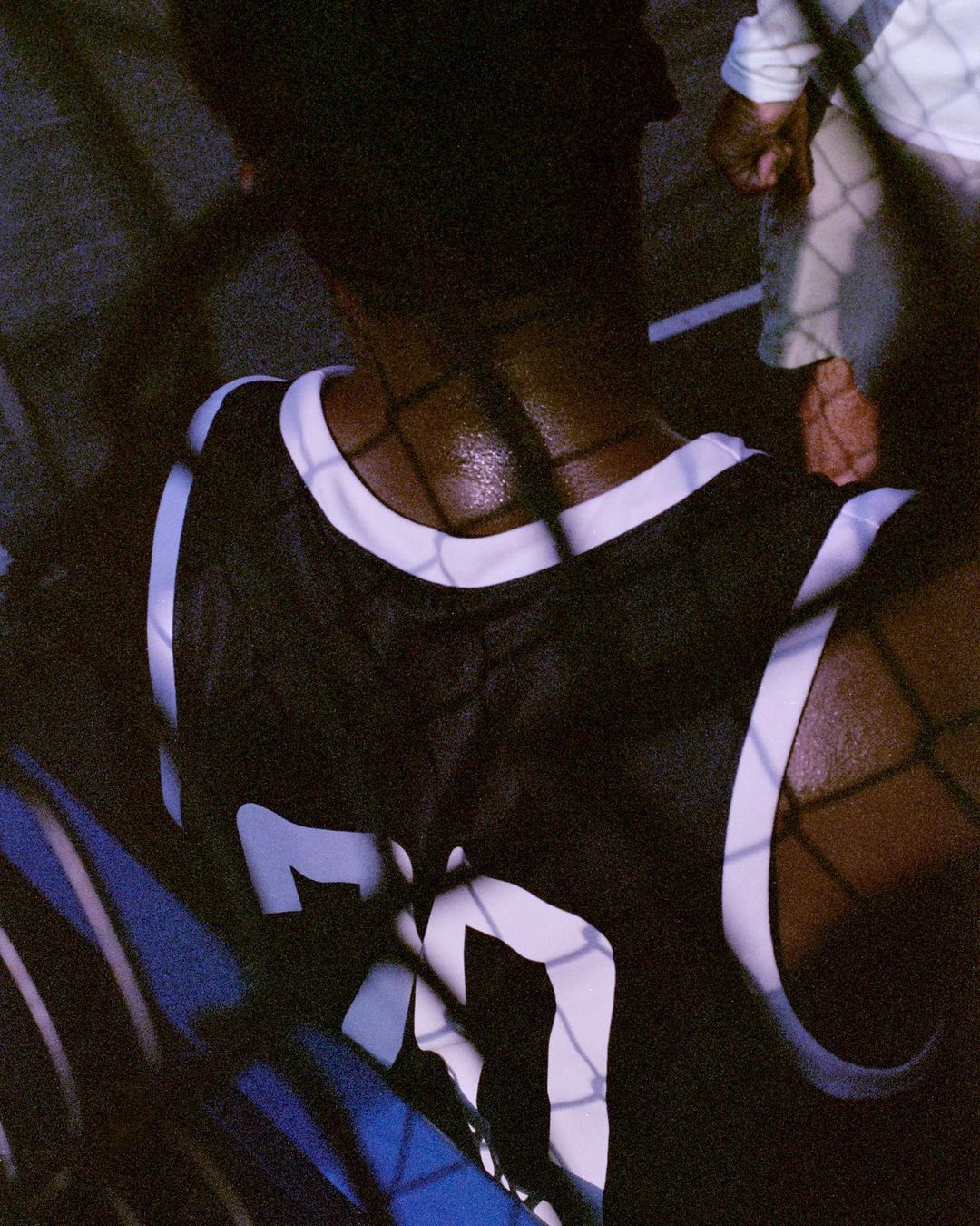 A person is seen from behind wearing a jersey with white trim, viewed through a wire fence in dim lighting, with a slight blue cast.