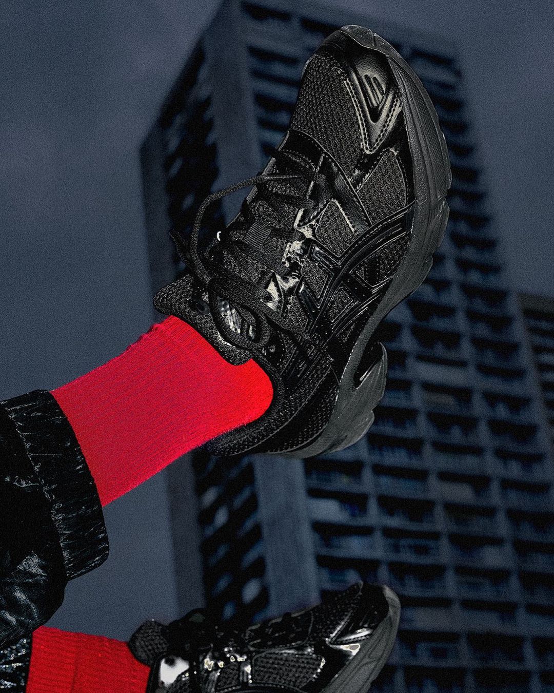 Elevated view of two black athletic shoes on feet with bright red socks, set against a backdrop of a tall, weathered building.