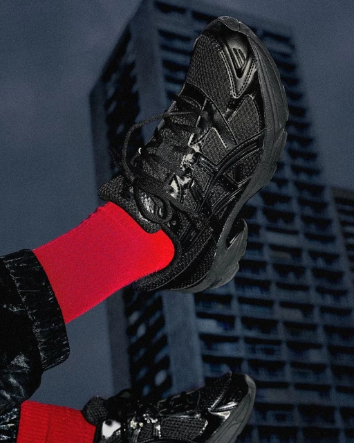 Elevated view of two black athletic shoes on feet with bright red socks, set against a backdrop of a tall, weathered building.