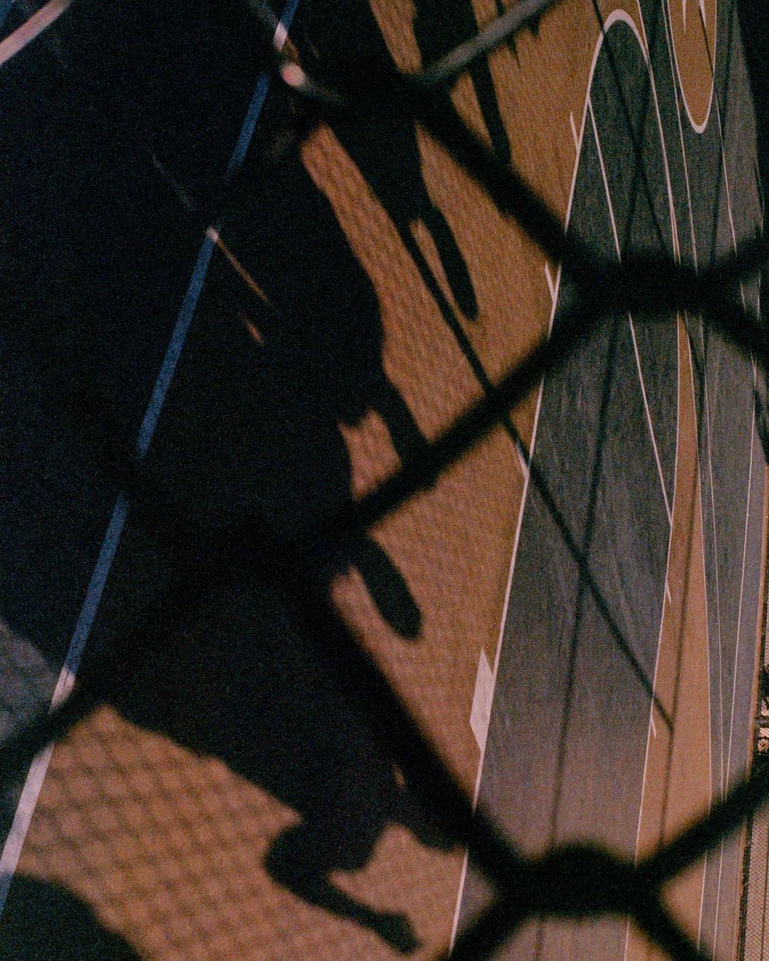 Nighttime shot of a dark basketball court seen through a chain-link fence, showcasing the court's lines and surface in dim lighting.