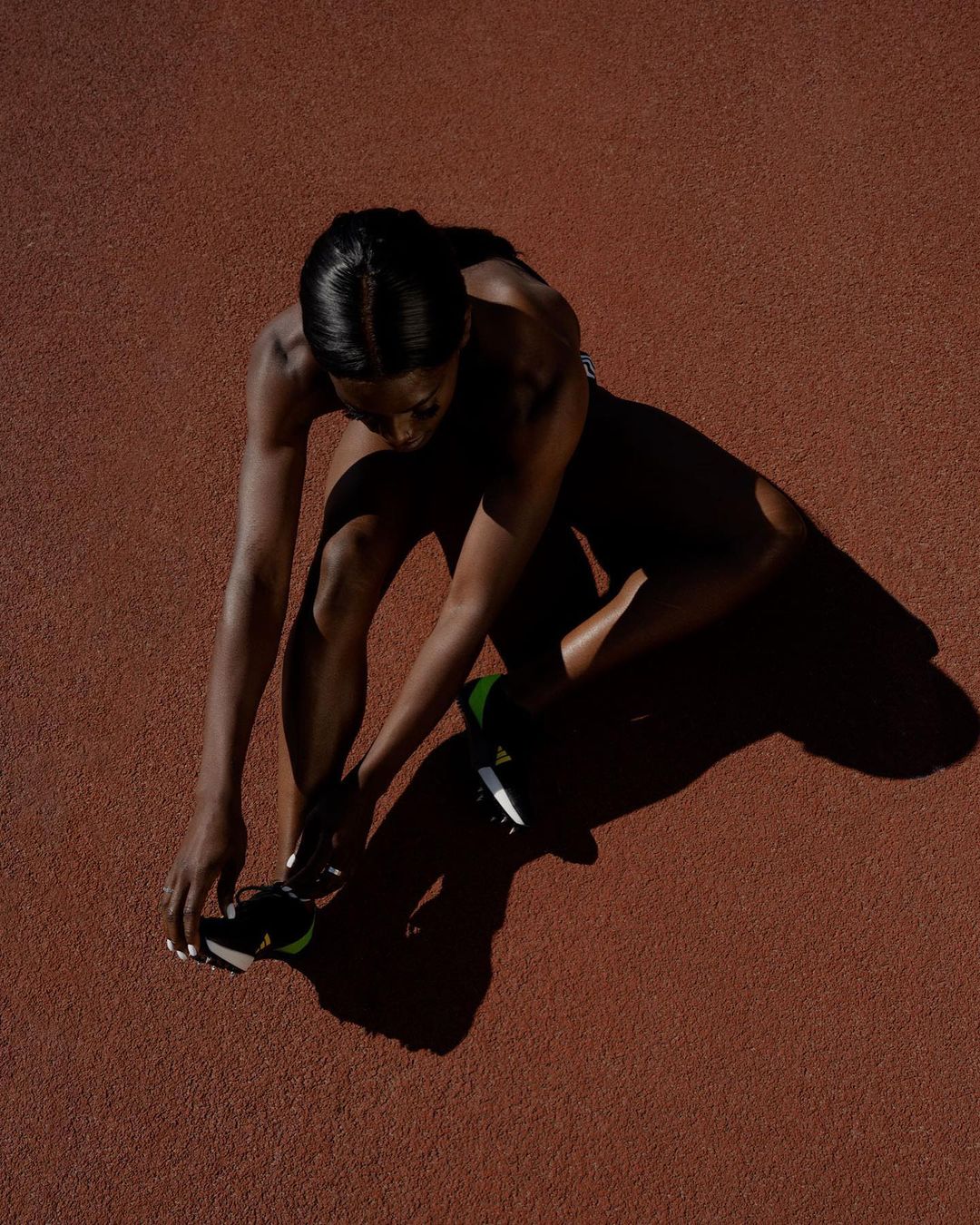 Overhead shot of a woman tying her running shoe on a red track, illuminated by bright sunlight casting long shadows.