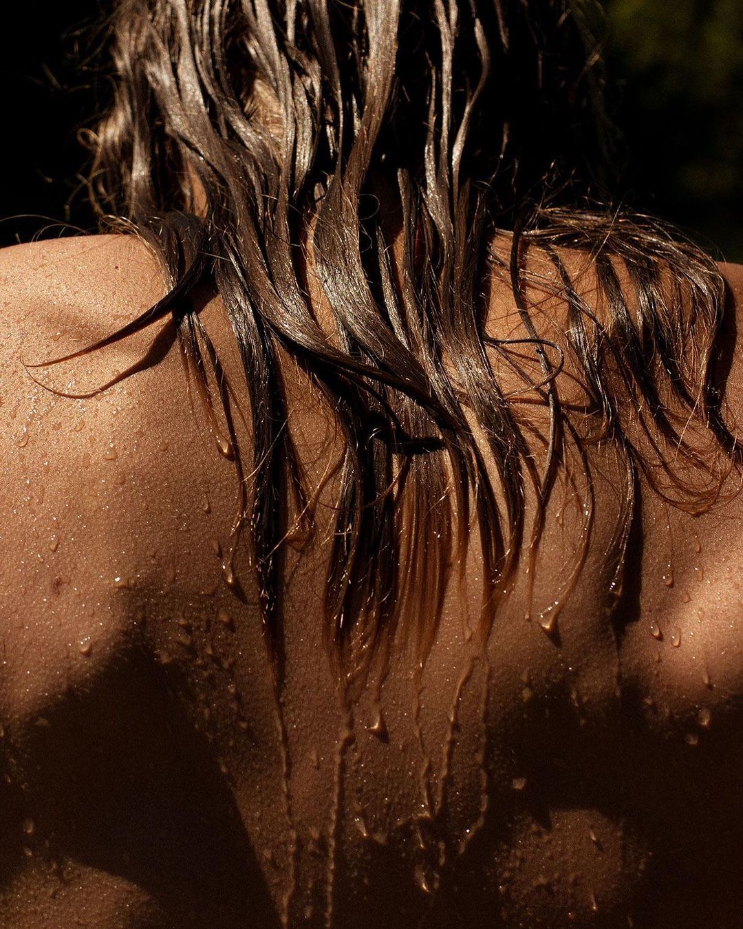 Close-up of the upper back and shoulders of a woman with wet hair cascading down her back, illuminated by dappled sunlight creating shadows.