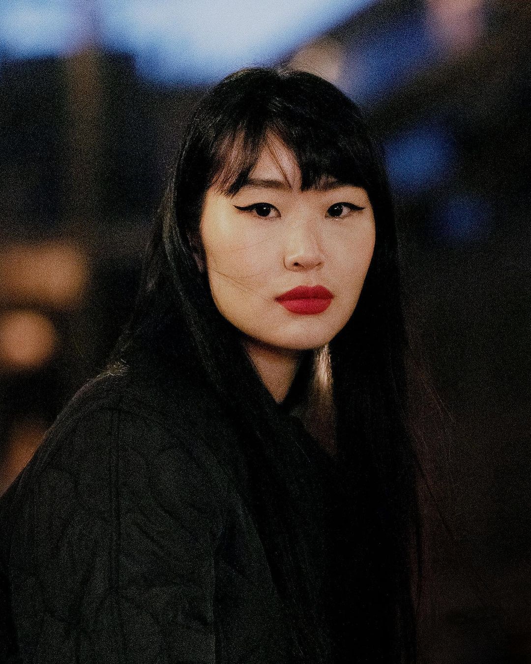 Portrait of an Asian woman with dark hair, red lipstick, wearing a dark jacket against a blurred background, showcasing a beauty shot.