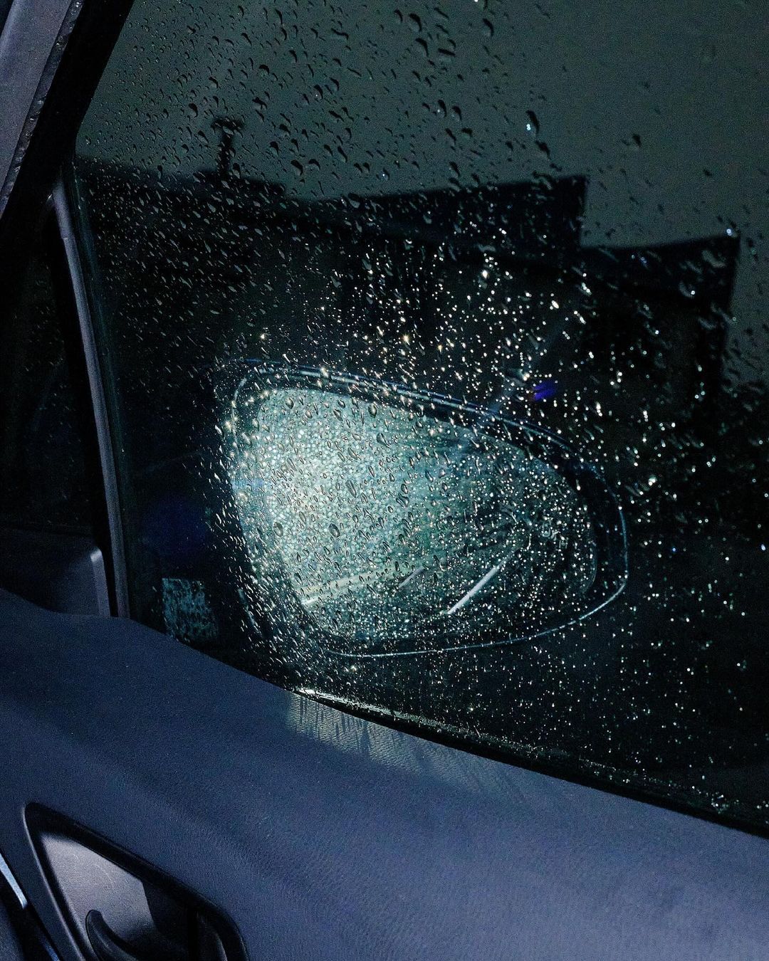 Raindrops cover a car window and side mirror, creating a blurred and wet view of the outside environment, under dark, overcast lighting.