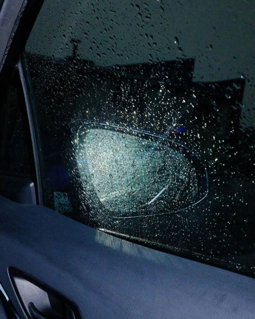 Raindrops cover a car window and side mirror, creating a blurred and wet view of the outside environment, under dark, overcast lighting.