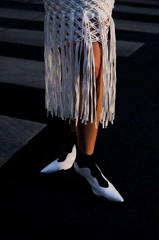 Woman wearing a short white fringe skirt and white pointed shoes walking on a street with zebra crossing lines in bright sunlight.