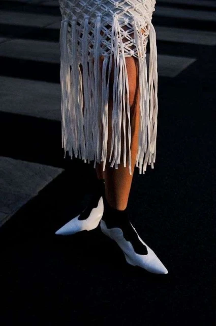 Woman wearing a short white fringe skirt and white pointed shoes walking on a street with zebra crossing lines in bright sunlight.