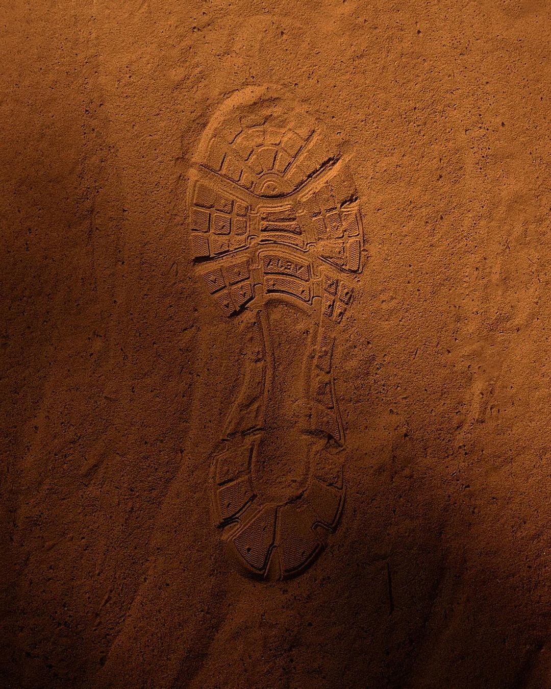 Close-up of a shoeprint in reddish-brown sand, lit from above, showing the sole's texture, shape, and slight depression.