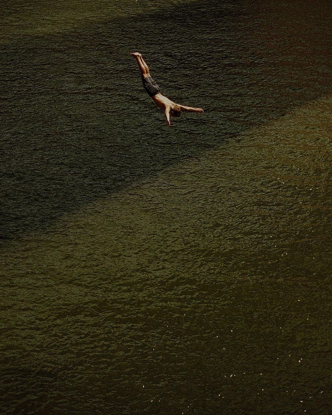 An adult male dives headfirst into dark water with arms outstretched in an outdoor environment on a sunny day from an eye-level vantage point.