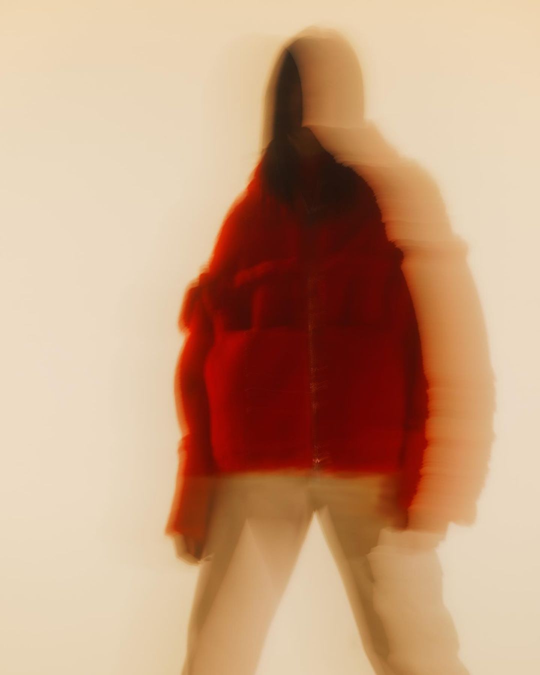 Blurred figure in motion wearing red jacket and light pants against a neutral backdrop, creating a sense of movement and abstraction.