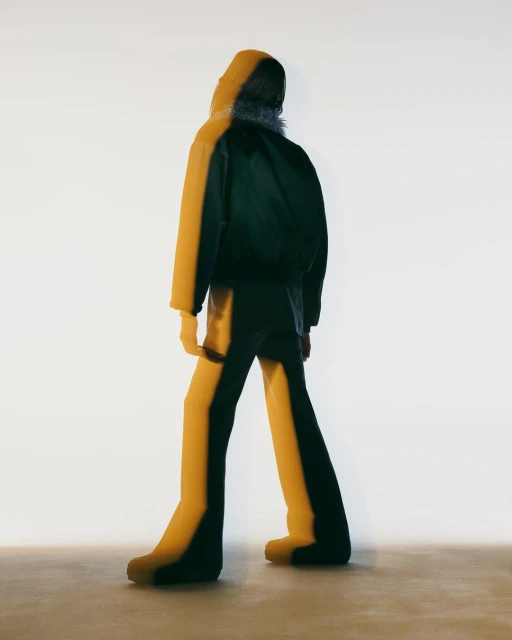 Back view of hooded figure in a dark jacket and pants, illuminated by a bright light with a faint double exposure effect. Studio shot.
