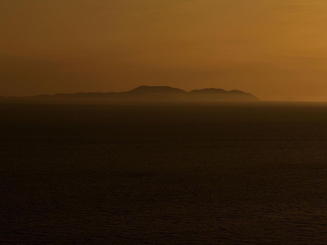 A view of a dark ocean and silhouetted islands under an orange sky during sunset, with dark shadows on the water.