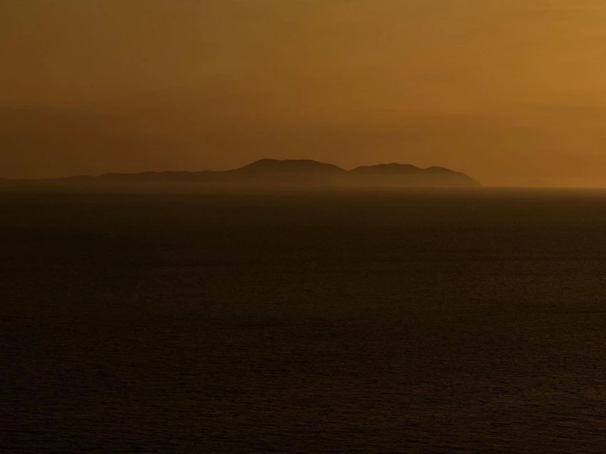 A view of a dark ocean and silhouetted islands under an orange sky during sunset, with dark shadows on the water.