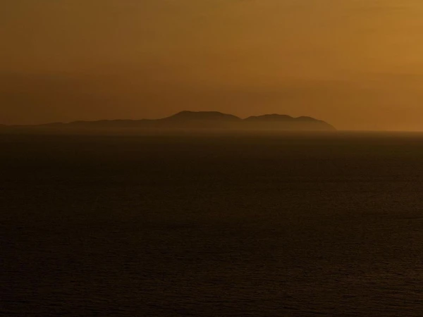 A view of a dark ocean and silhouetted islands under an orange sky during sunset, with dark shadows on the water.