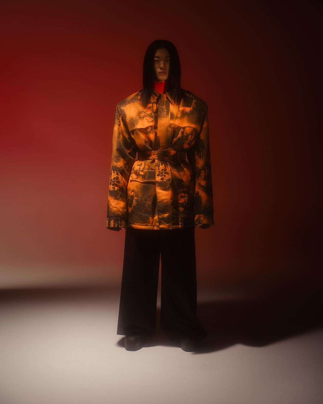 Full-length portrait of a stylish Asian male model with long hair wearing a patterned jacket and wide black pants against red backdrop.