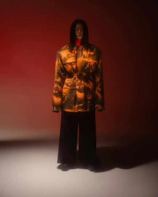 Full-length portrait of a stylish Asian male model with long hair wearing a patterned jacket and wide black pants against red backdrop.