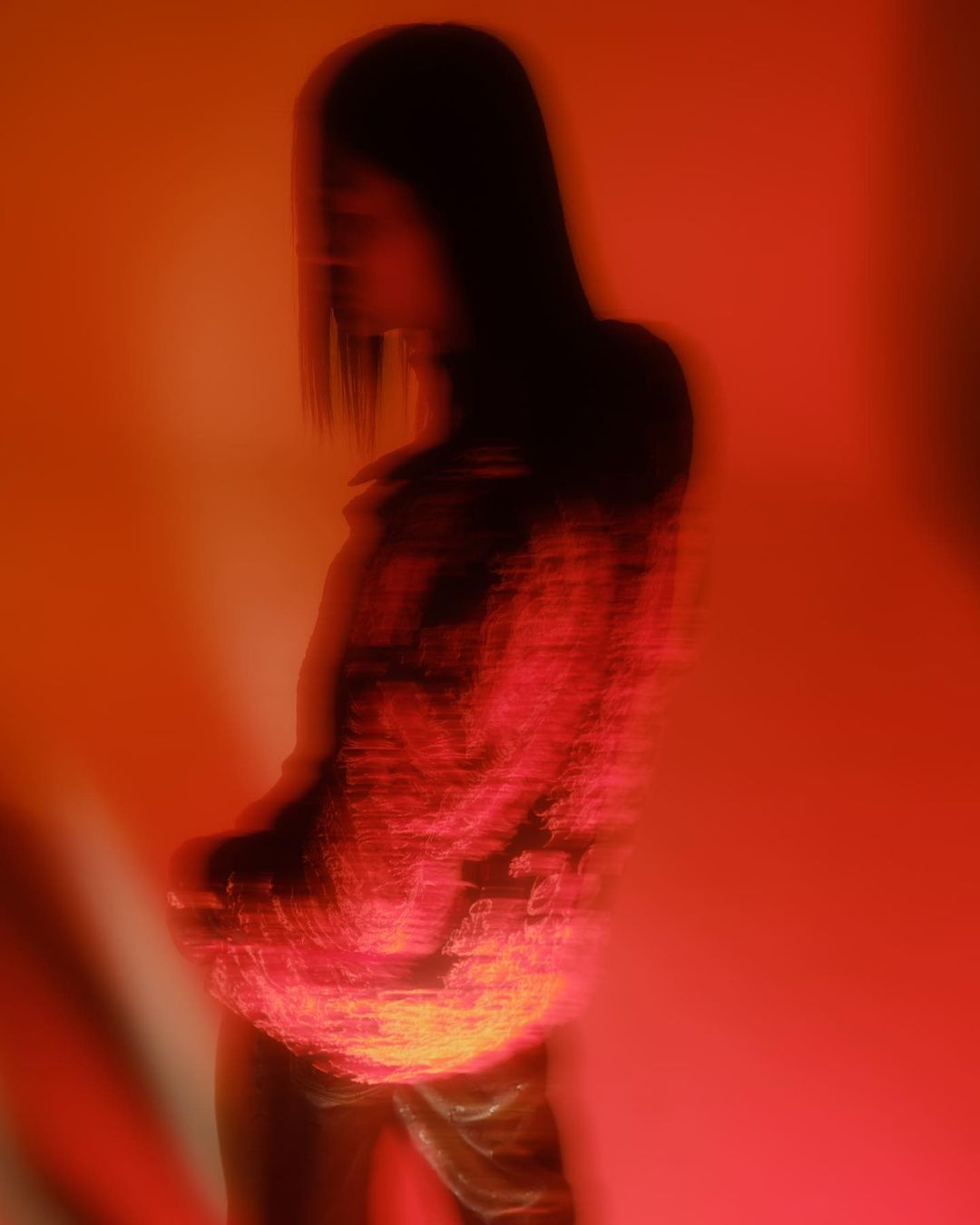 Abstract portrait of a blurry woman with long dark hair posing in an orange-lit photo studio wearing a patterned red top.