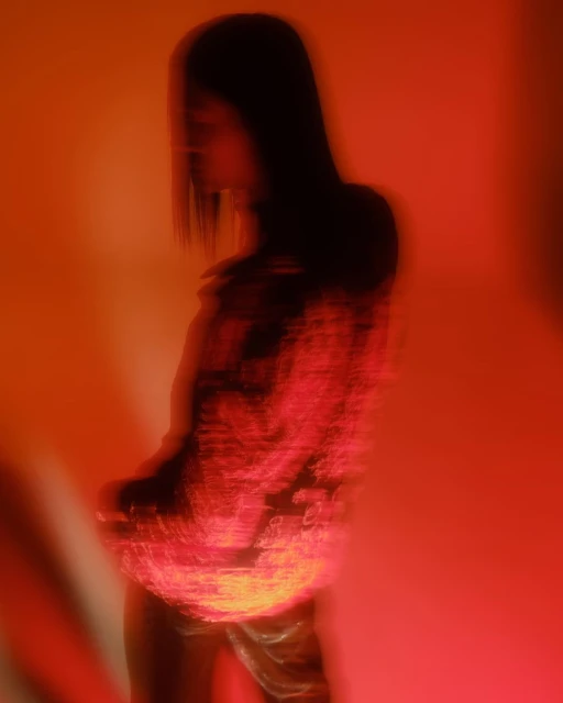 Abstract portrait of a blurry woman with long dark hair posing in an orange-lit photo studio wearing a patterned red top.