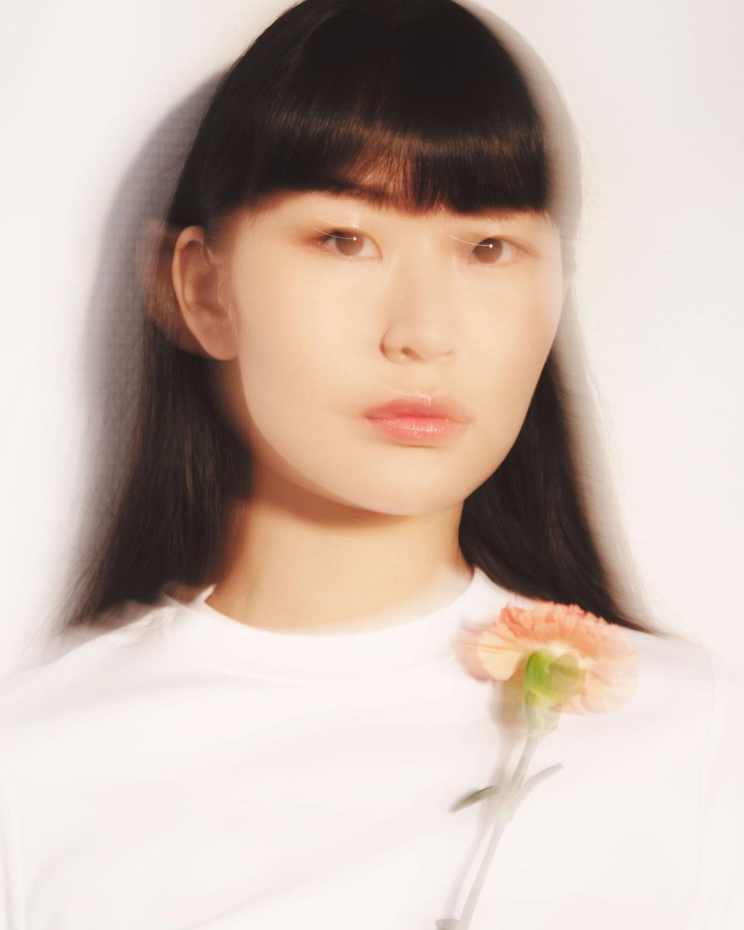 Portrait of an Asian woman with dark hair wearing a white t-shirt with a light orange carnation flower pinned to her chest in a photo studio.