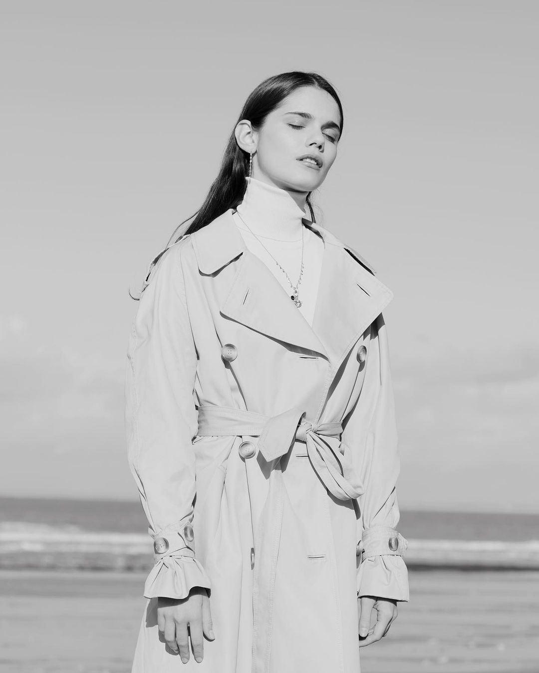 Monochrome shot of a young woman with eyes closed wearing a trench coat on a beach under a clear sky, capturing a serene outdoor fashion moment.