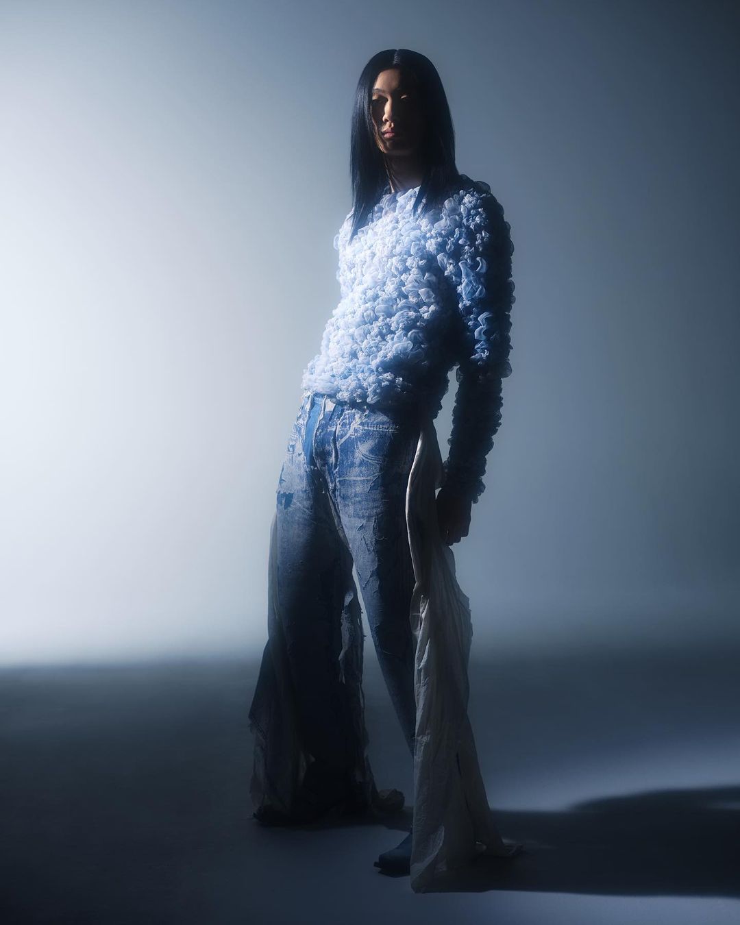 Full-length shot of a slender person with long black hair wearing a blue knit top and baggy, distressed jeans in a studio setting.
