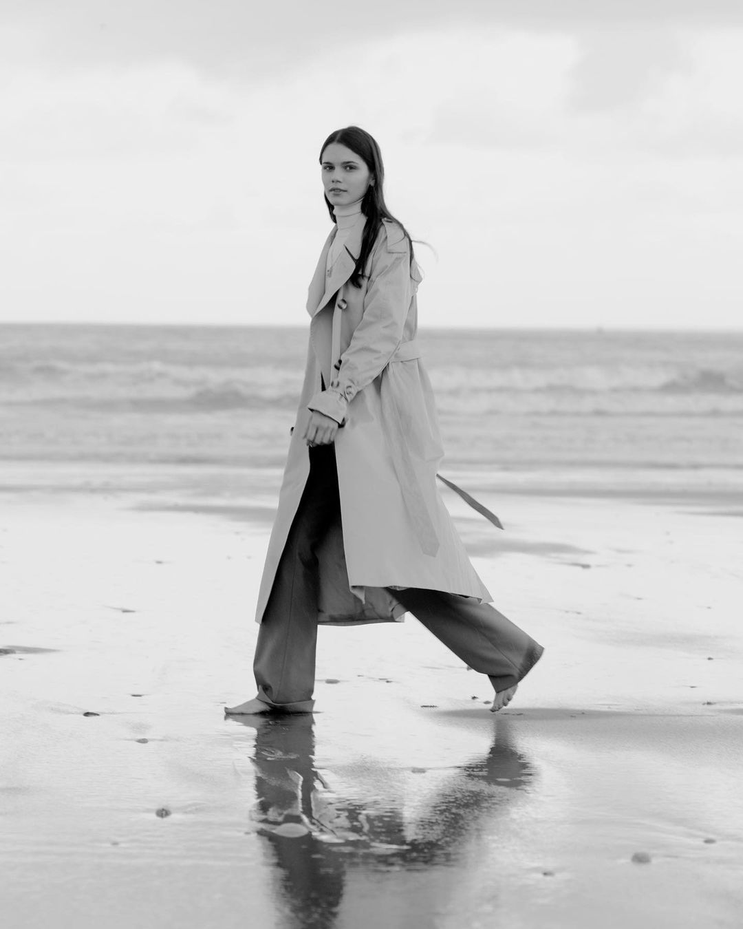 Monochrome portrait of a barefoot adult female walking on the beach, wearing a trench coat with the ocean and sky in the background.