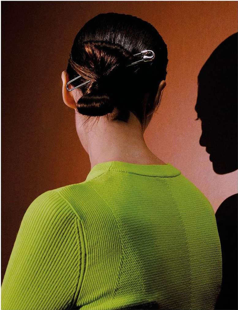 Back of a woman with dark hair styled in a bun and secured with silver safety pins, wearing a bright green ribbed top, against a brown backdrop.