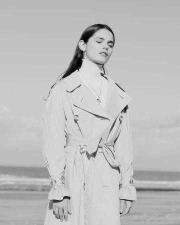 Black and white portrait of a woman wearing a belted trench coat and turtleneck outdoors by the ocean, photographed by Anne-Sophie Soudoplatoff.