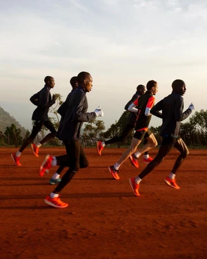 Group of runners in dark jackets and bright orange running shoes training on a red dirt track under an open sky.