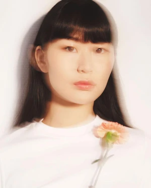 Portrait of a woman with long dark hair wearing a white t-shirt adorned with a single peach flower against a bright white background.