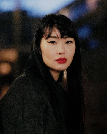 Portrait of an Asian woman with long black hair, winged eyeliner, and red lipstick wearing a dark quilted jacket.