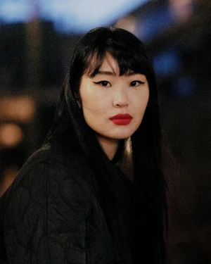 Portrait of an Asian woman with long black hair, winged eyeliner, and red lipstick wearing a dark quilted jacket.