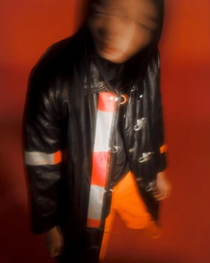 Motion blurred portrait of a person in a black leather coat and bright orange trousers against a red background by Anne-Sophie Soudoplatoff.