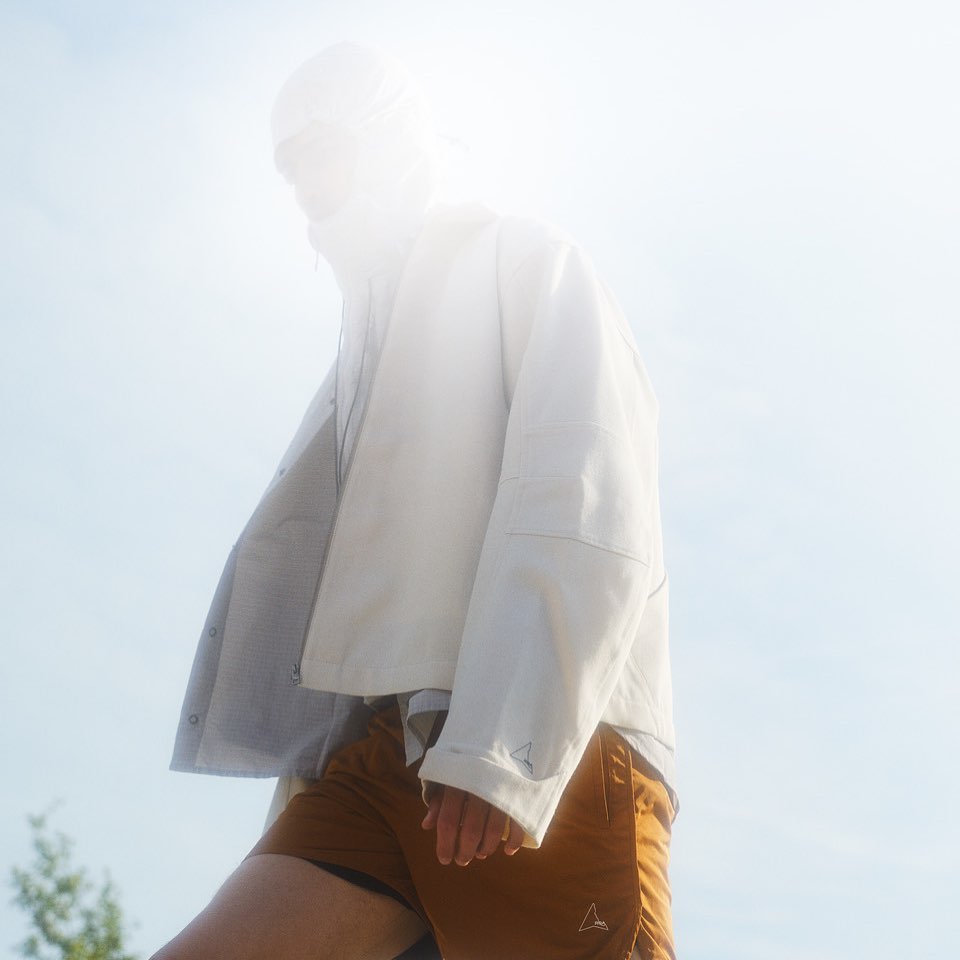 Low-angle shot of a model wearing a white jacket and brown shorts against a bright sky background.
