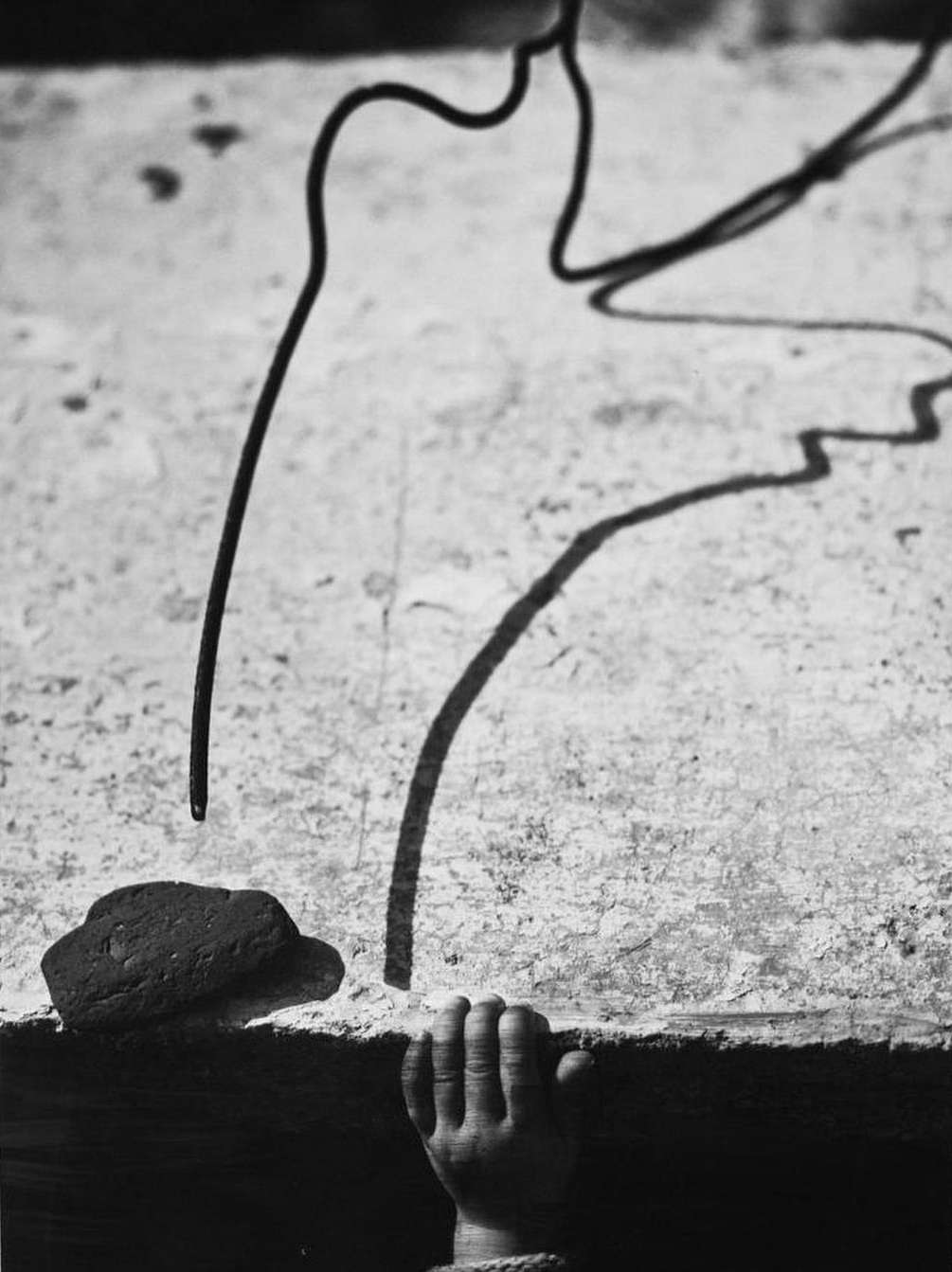 Black and white photography of a child's hand reaching up to a textured surface with a wire and brick, creating shadow play.