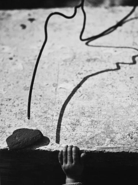 Black and white photography of a child's hand reaching up to a textured surface with a wire and brick, creating shadow play.